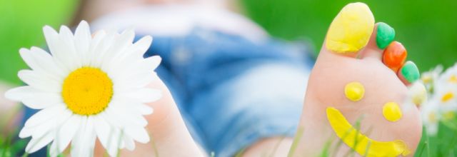 nurmikolla lepäävät jalkapohjat: toisessa jalassa kiinni kukka ja toisessa jalassa maalatut varpaat ja hymynaama