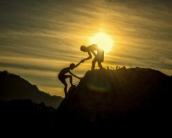 Ihminen auttaa toista pääsemään vuoren huipulle auringonlaskun alla