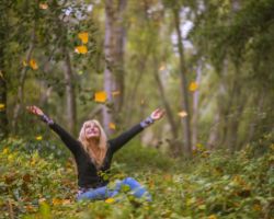 Nainen seisoo keskellä metsää ja heittää iloisena puunlehtiä ympärilleen