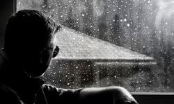 Aurinkolasipäinen mies katsoo ulos ikkunasta sateista säätä.