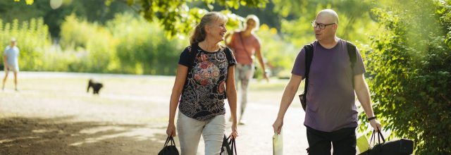 Mies ja nainen kävelevät yhdessä kesäisessä puistossa iloisesti jutellen, kantaen tavaroita käsissään.