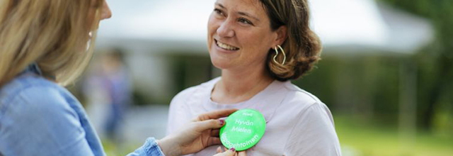 Nainen kiinnittää toisen hymyilevän rintaan ison rintamerkin, jossa lukee Hyvän mielen vapaaehtoinen